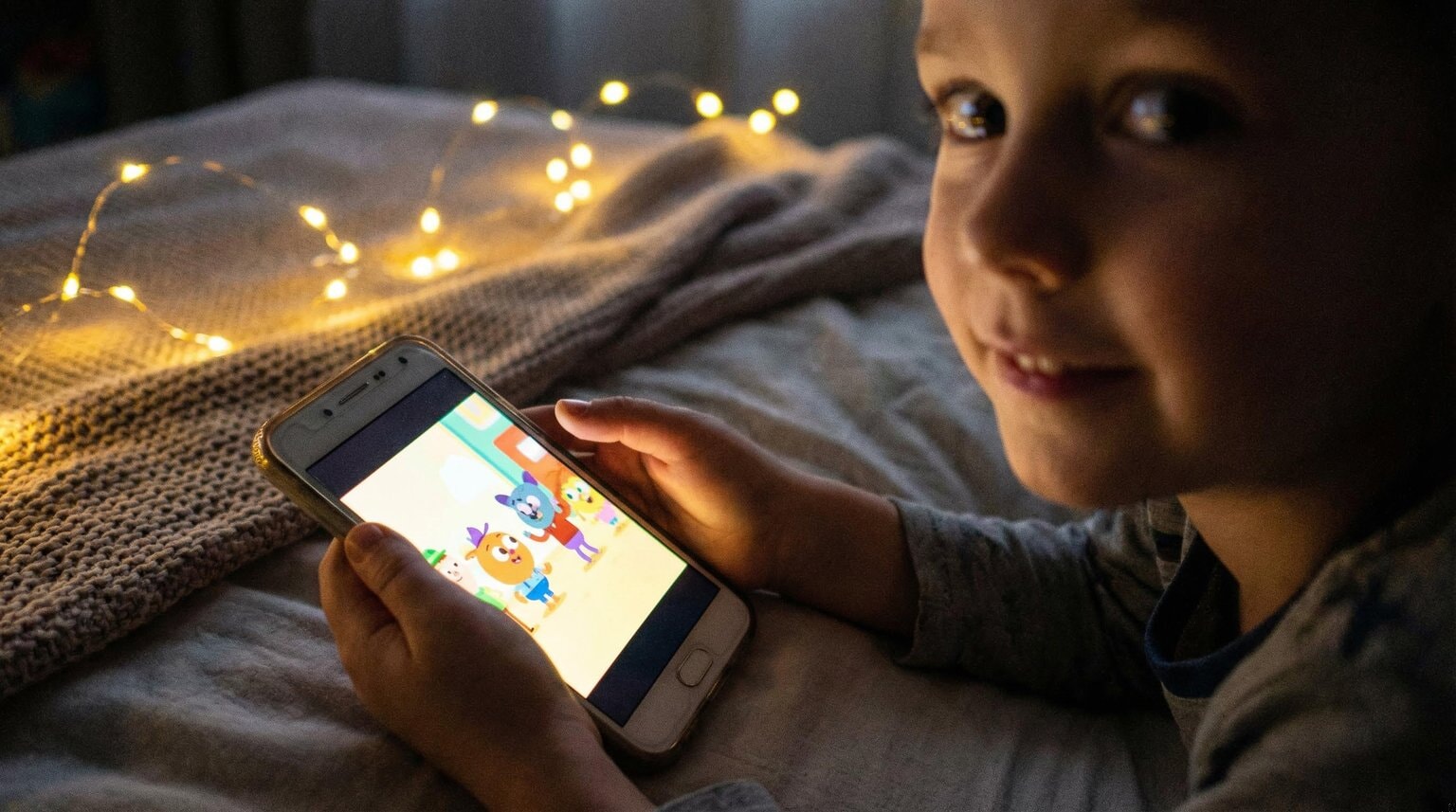 Child's hands holding smartphone with colorful video content glowing in dimly lit bedroom with fairy lights